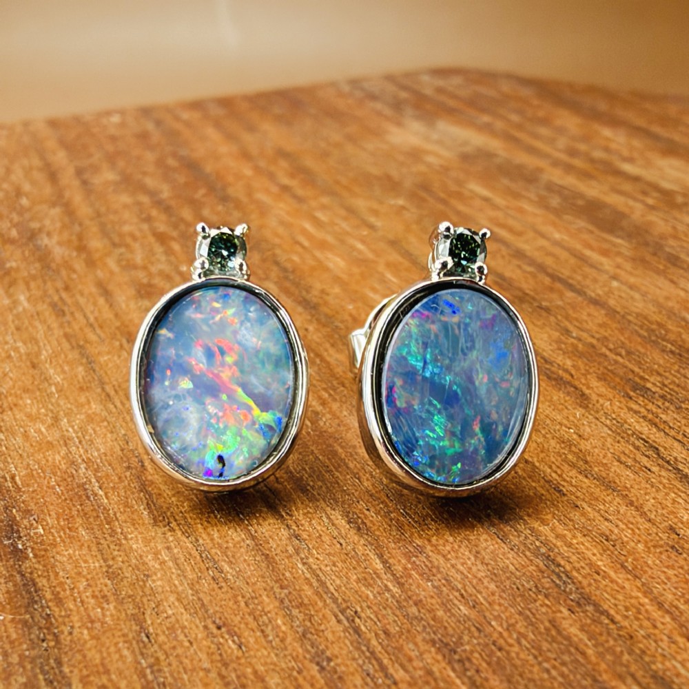 A pair of 14ct white gold stud earrings featuring oval opal doublets with blue and green flashes, each crowned with a small round prong-set teal green diamond. View IX