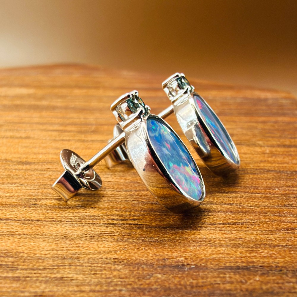 A pair of 14ct white gold stud earrings featuring oval opal doublets with blue and green flashes, each crowned with a small round prong-set teal green diamond. View X