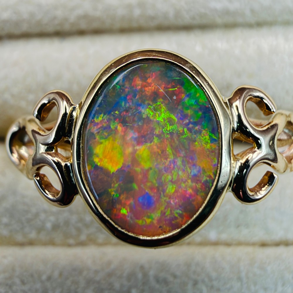 An 18ct gold ring featuring a central oval bezel-set opal displaying colourful flash, flanked by openwork scrolled shoulders. View V