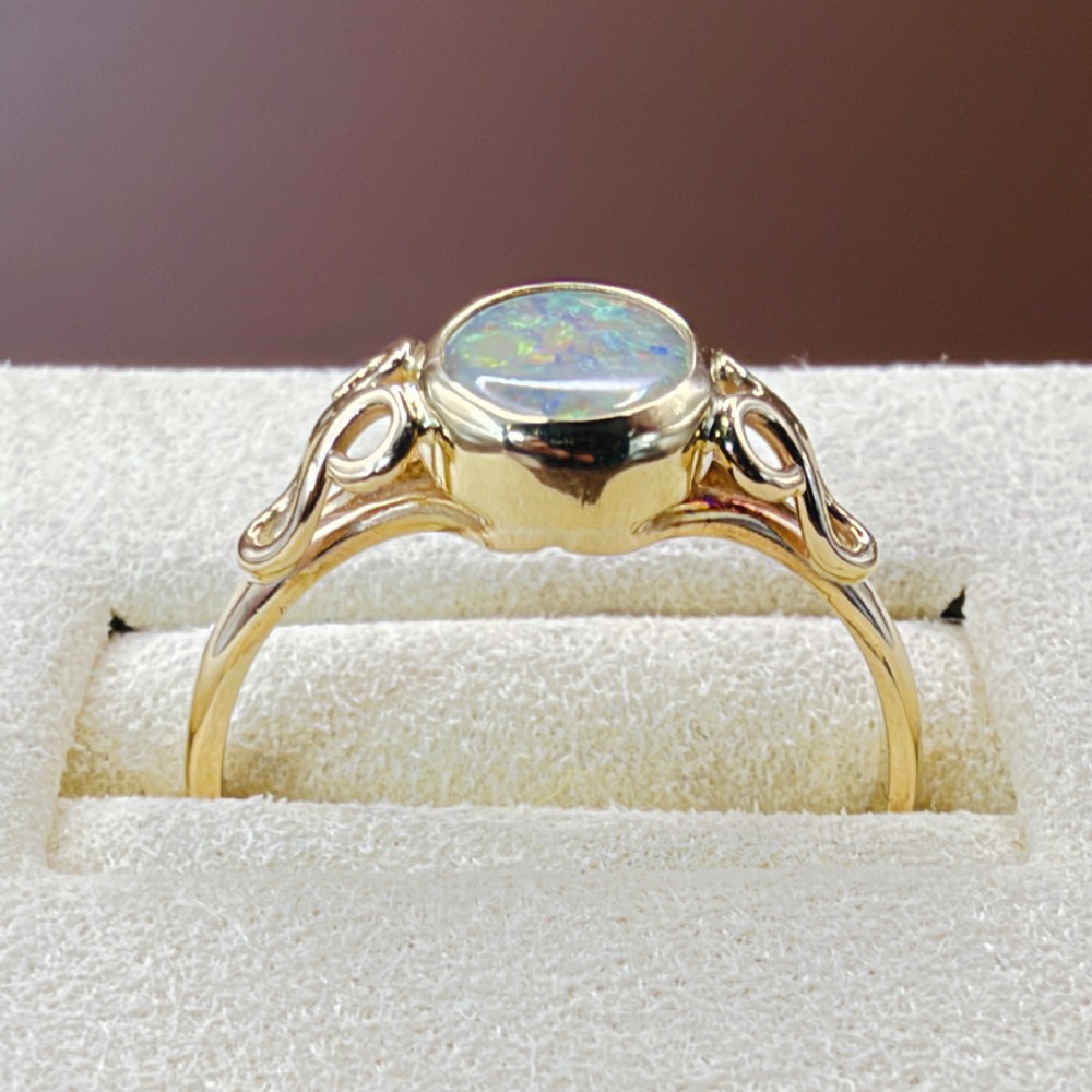 An 18ct gold ring featuring a central oval bezel-set opal displaying colourful flash, flanked by openwork scrolled shoulders. View VII