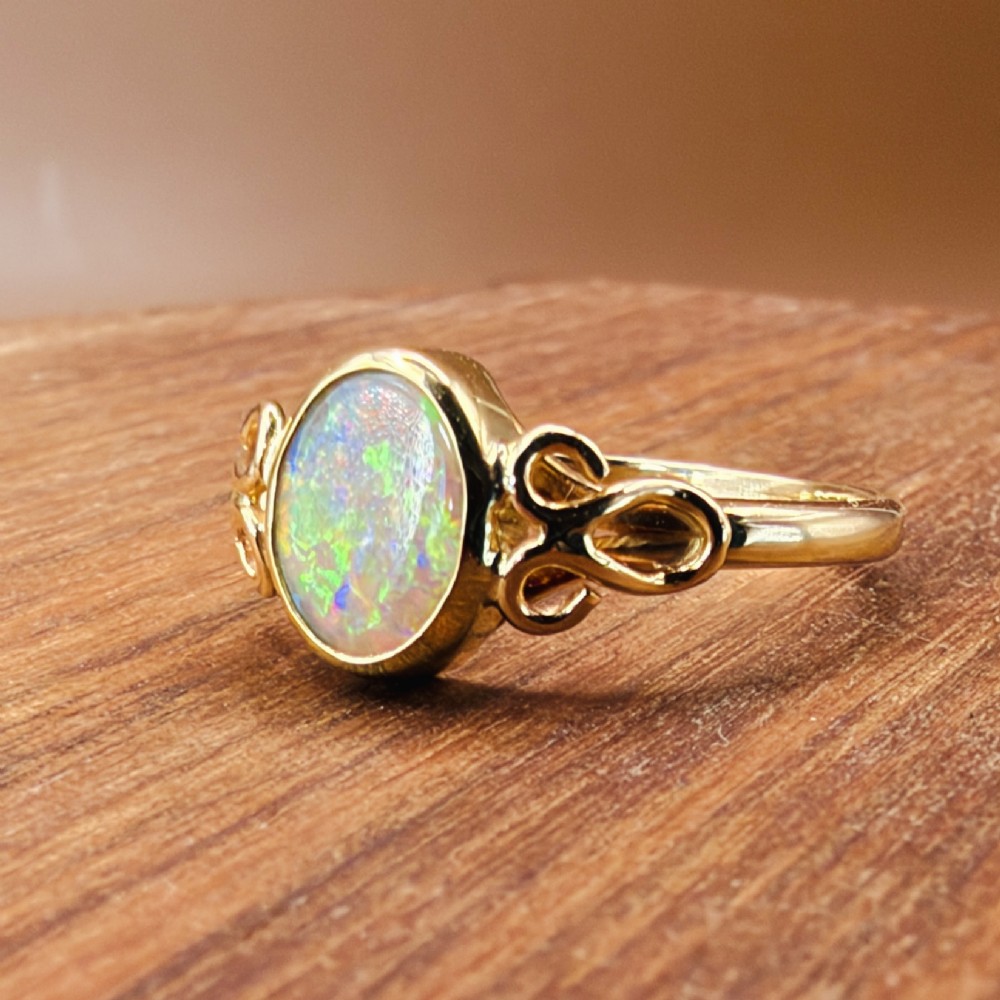 An 18ct gold ring featuring a central oval bezel-set opal displaying colourful flash, flanked by openwork scrolled shoulders. View VIII