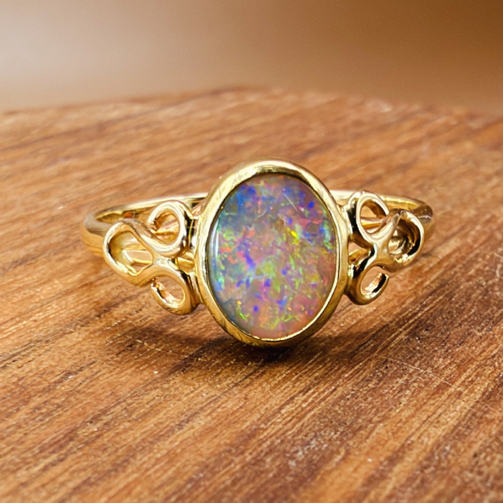 An 18ct gold ring featuring a central oval bezel-set opal displaying colourful flash, flanked by openwork scrolled shoulders. View IX