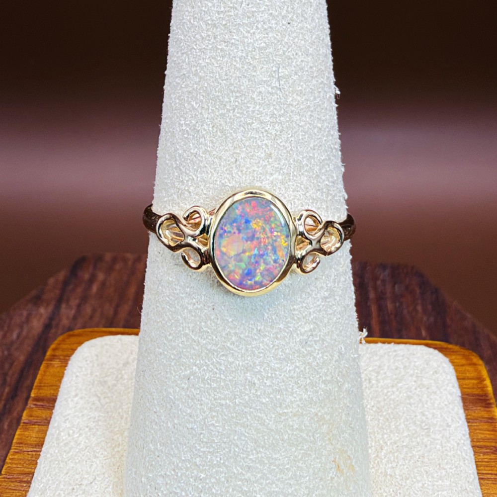 An 18ct gold ring featuring a central oval bezel-set opal displaying colourful flash, flanked by openwork scrolled shoulders. View X