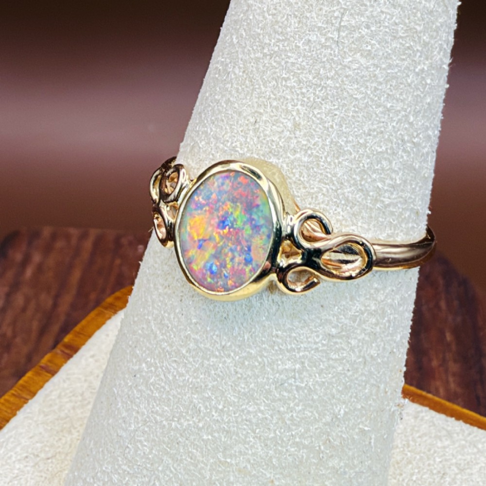 An 18ct gold ring featuring a central oval bezel-set opal displaying colourful flash, flanked by openwork scrolled shoulders. View XI