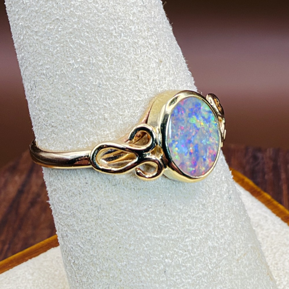 An 18ct gold ring featuring a central oval bezel-set opal displaying colourful flash, flanked by openwork scrolled shoulders. View XII
