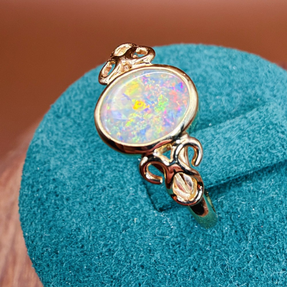An 18ct gold ring featuring a central oval bezel-set opal displaying colourful flash, flanked by openwork scrolled shoulders. View XIII