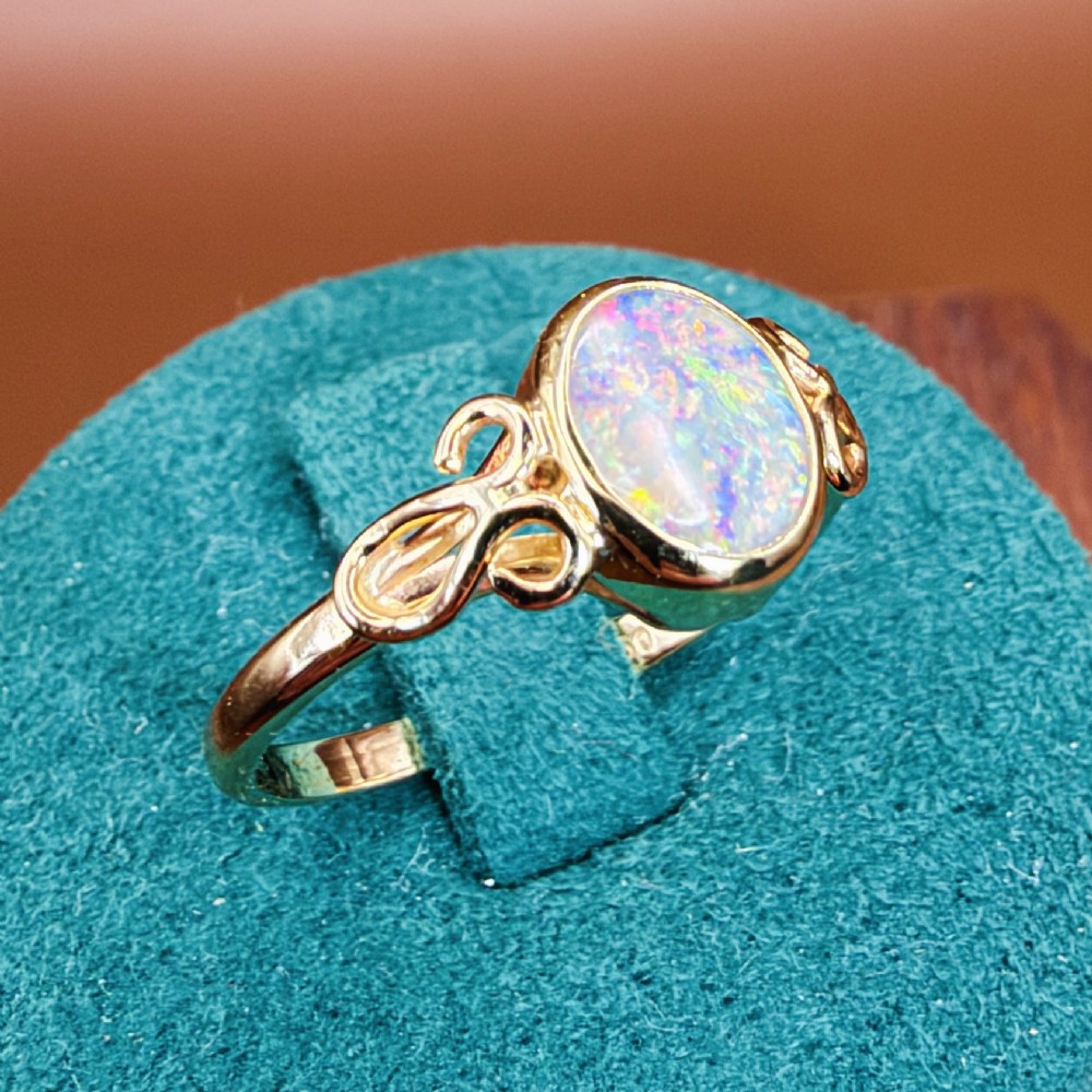 An 18ct gold ring featuring a central oval bezel-set opal displaying colourful flash, flanked by openwork scrolled shoulders. View XIV