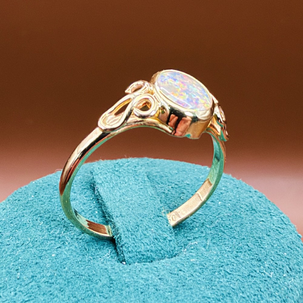 An 18ct gold ring featuring a central oval bezel-set opal displaying colourful flash, flanked by openwork scrolled shoulders. View XV