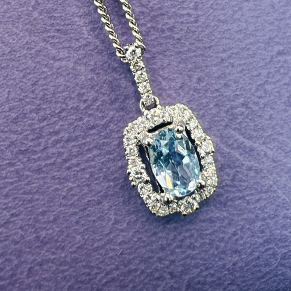 An 18ct white gold pendant featuring a central oval-cut light blue aquamarine surrounded by a frame of round brilliant diamonds with a diamond-set bail. View IV
