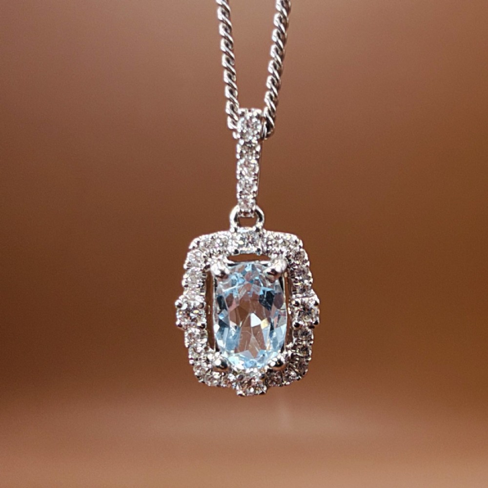 An 18ct white gold pendant featuring a central oval-cut light blue aquamarine surrounded by a frame of round brilliant diamonds with a diamond-set bail. View V