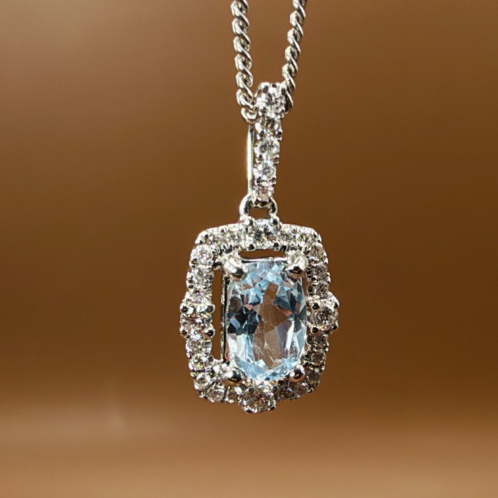 An 18ct white gold pendant featuring a central oval-cut light blue aquamarine surrounded by a frame of round brilliant diamonds with a diamond-set bail. View VI