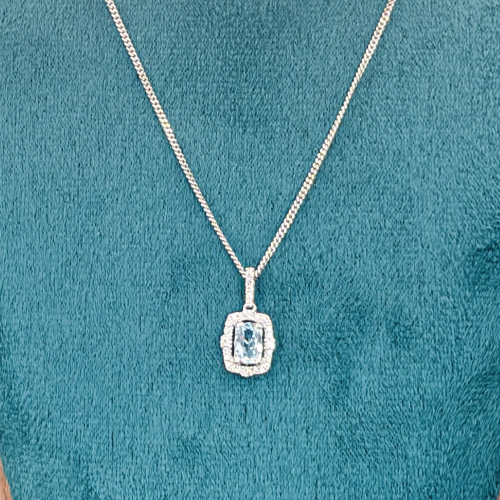 An 18ct white gold pendant featuring a central oval-cut light blue aquamarine surrounded by a frame of round brilliant diamonds with a diamond-set bail. View X