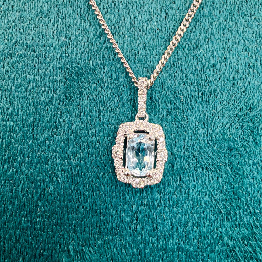 An 18ct white gold pendant featuring a central oval-cut light blue aquamarine surrounded by a frame of round brilliant diamonds with a diamond-set bail. View XI