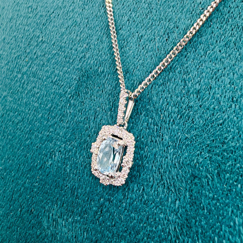An 18ct white gold pendant featuring a central oval-cut light blue aquamarine surrounded by a frame of round brilliant diamonds with a diamond-set bail. View XII