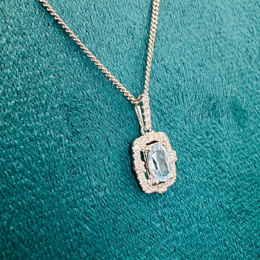 An 18ct white gold pendant featuring a central oval-cut light blue aquamarine surrounded by a frame of round brilliant diamonds with a diamond-set bail. View XIII