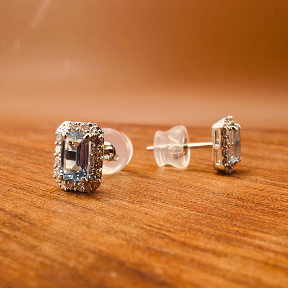 A pair of platinum stud earrings featuring emerald-cut light blue aquamarines surrounded by a border of brilliant-cut diamonds. View VI