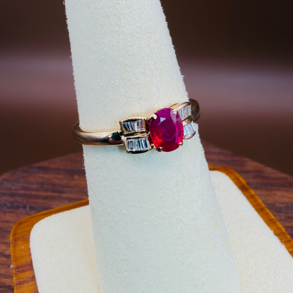 An 18ct yellow gold ring featuring an oval cut central red ruby flanked by stepped baguette cut diamonds on a polished band. View VIII