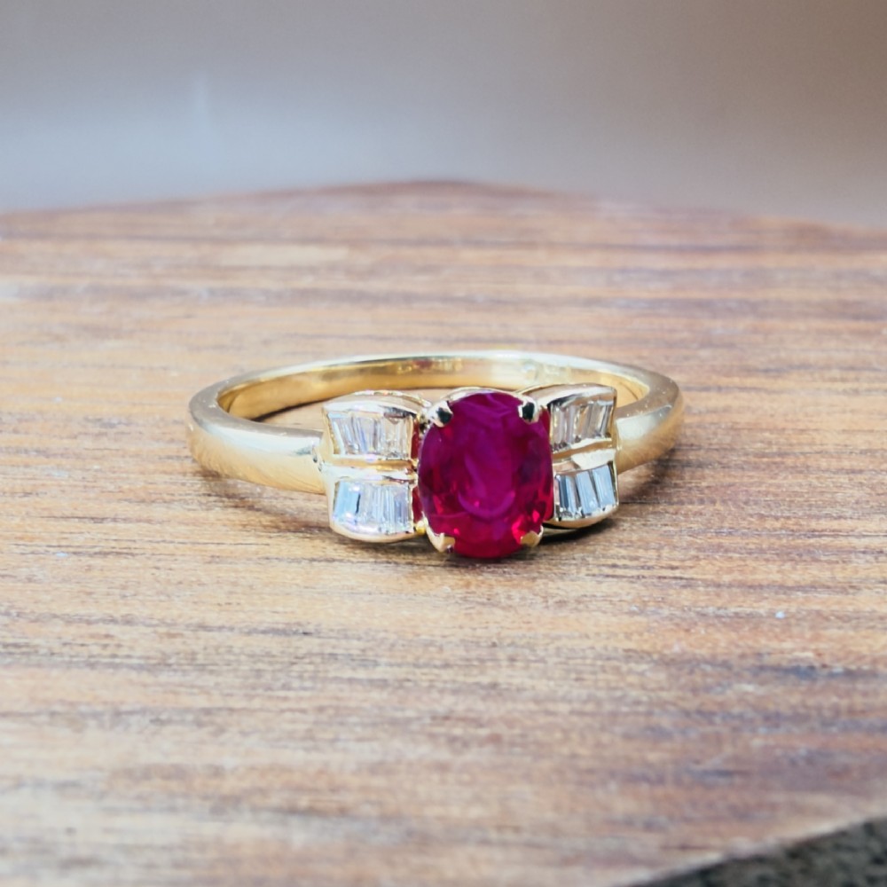 An 18ct yellow gold ring featuring an oval cut central red ruby flanked by stepped baguette cut diamonds on a polished band. View X