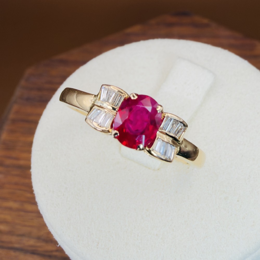 An 18ct yellow gold ring featuring an oval cut central red ruby flanked by stepped baguette cut diamonds on a polished band.