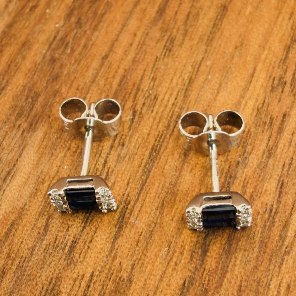 A pair of 9ct white gold stud earrings featuring two central emerald-cut blue sapphires flanked by small round diamonds. View V