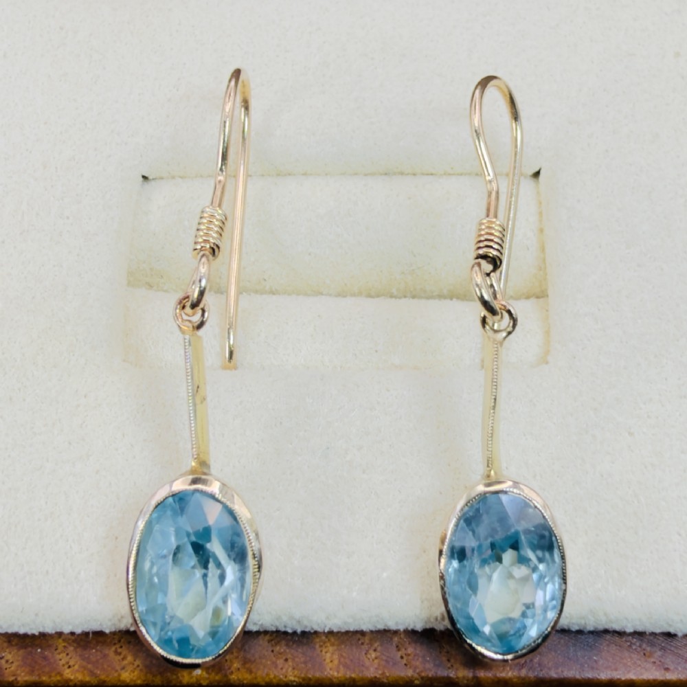 A pair of 9ct yellow gold Art Deco drop earrings featuring oval-cut light blue zircons suspended from vertical gold bars and shepherd hook wires.
