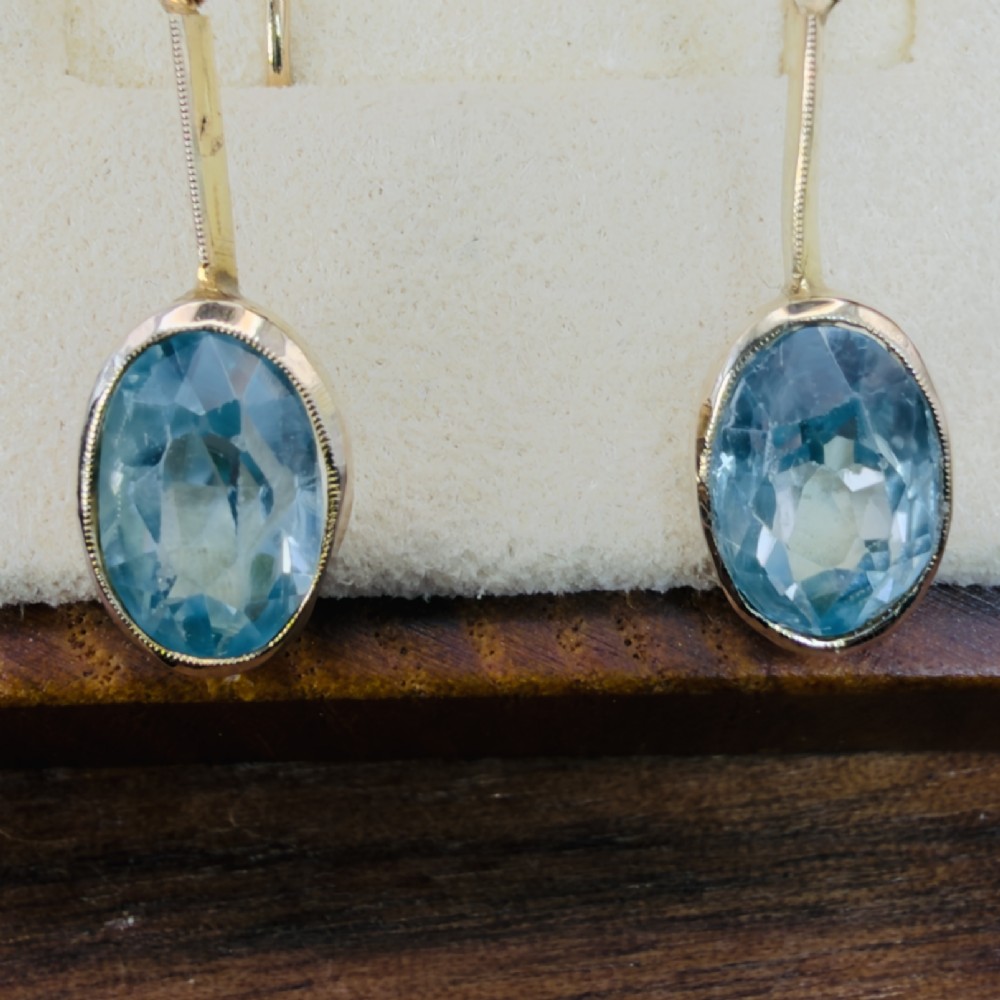 A pair of 9ct yellow gold Art Deco drop earrings featuring oval-cut light blue zircons suspended from vertical gold bars and shepherd hook wires. View IV