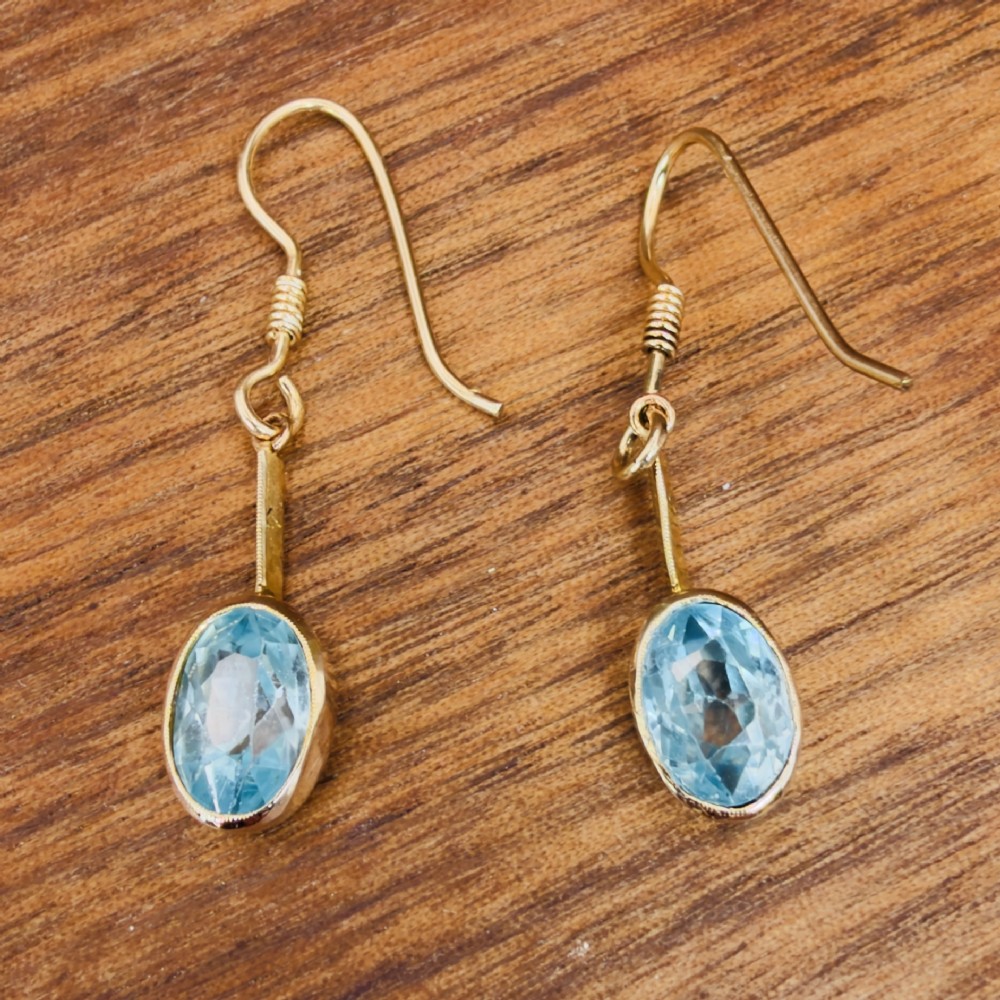 A pair of 9ct yellow gold Art Deco drop earrings featuring oval-cut light blue zircons suspended from vertical gold bars and shepherd hook wires. View X