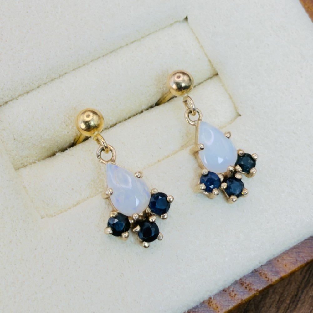 A pair of 9ct yellow gold stud drop earrings featuring pear-shaped white opal cabochons accented with three round blue sapphires at the base. View IV