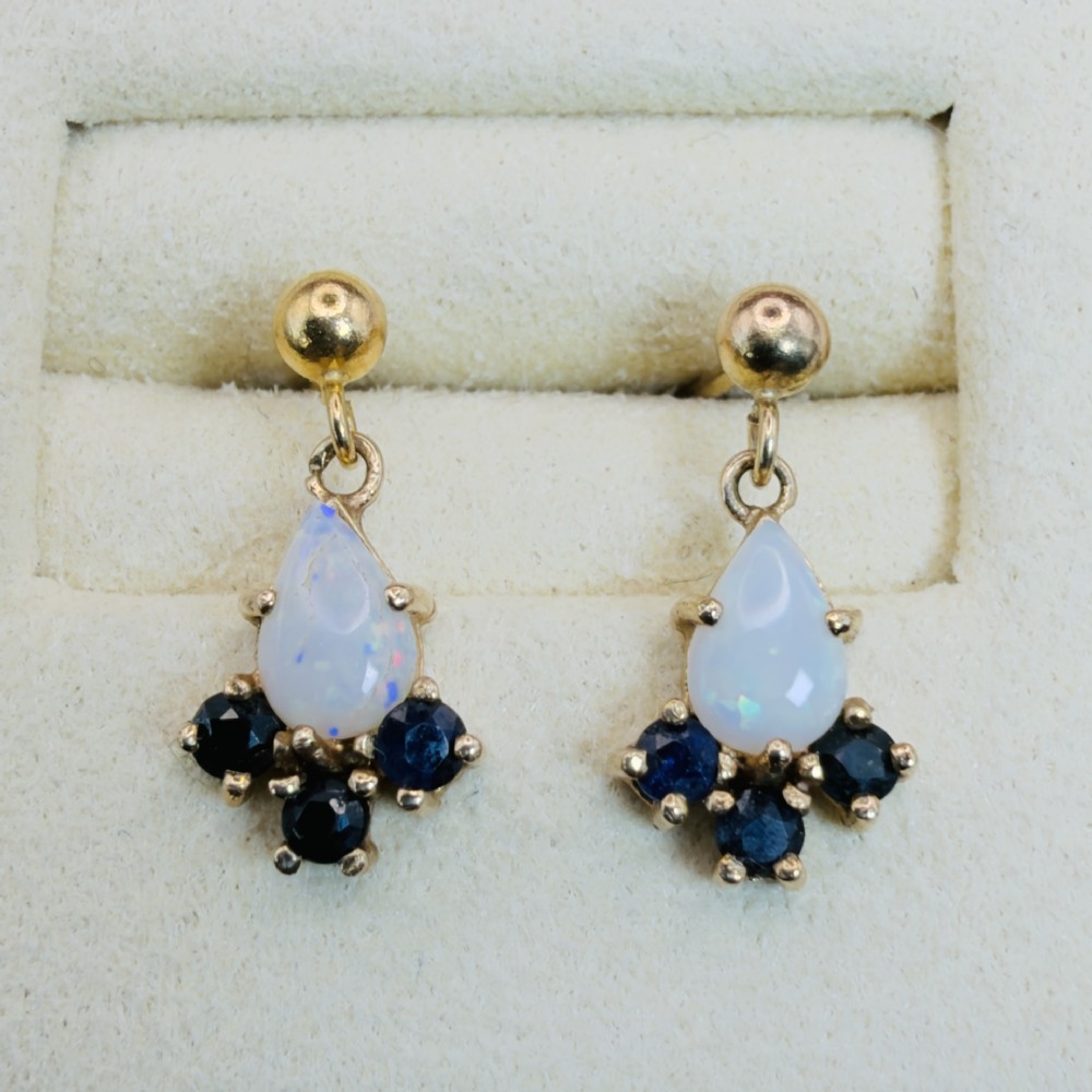 A pair of 9ct yellow gold stud drop earrings featuring pear-shaped white opal cabochons accented with three round blue sapphires at the base.