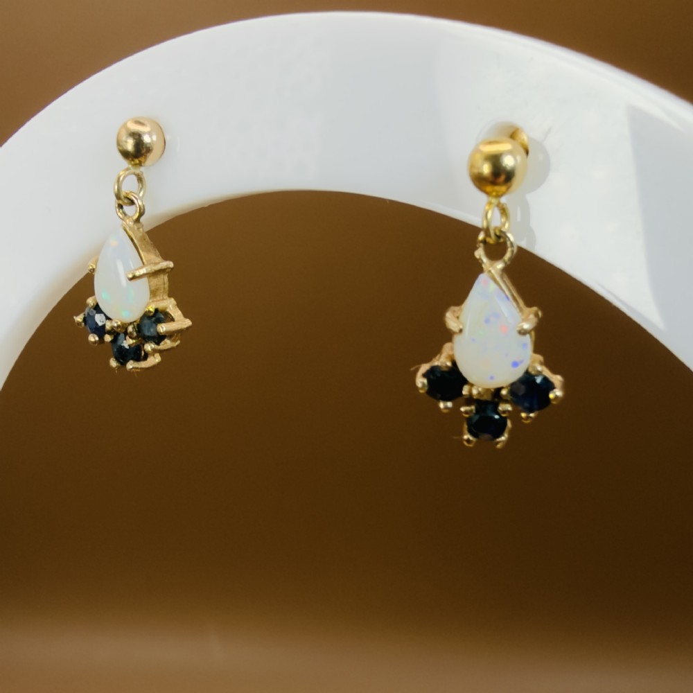 A pair of 9ct yellow gold stud drop earrings featuring pear-shaped white opal cabochons accented with three round blue sapphires at the base. View VI