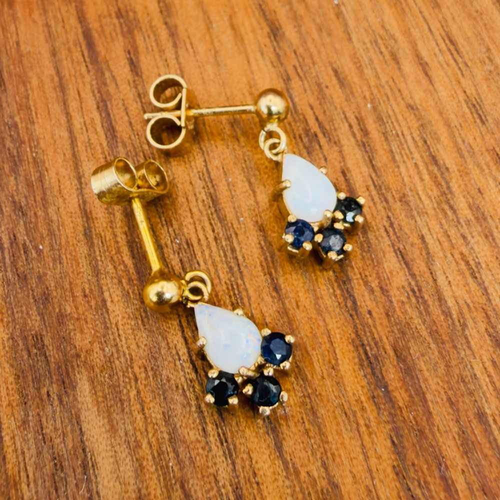A pair of 9ct yellow gold stud drop earrings featuring pear-shaped white opal cabochons accented with three round blue sapphires at the base. View IX