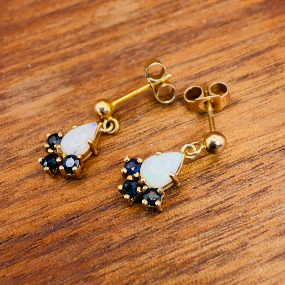 A pair of 9ct yellow gold stud drop earrings featuring pear-shaped white opal cabochons accented with three round blue sapphires at the base. View X