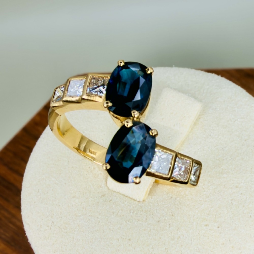 An 18ct yellow gold bypass ring featuring two dark blue oval-cut sapphires accented by channel-set princess-cut diamonds along the band.