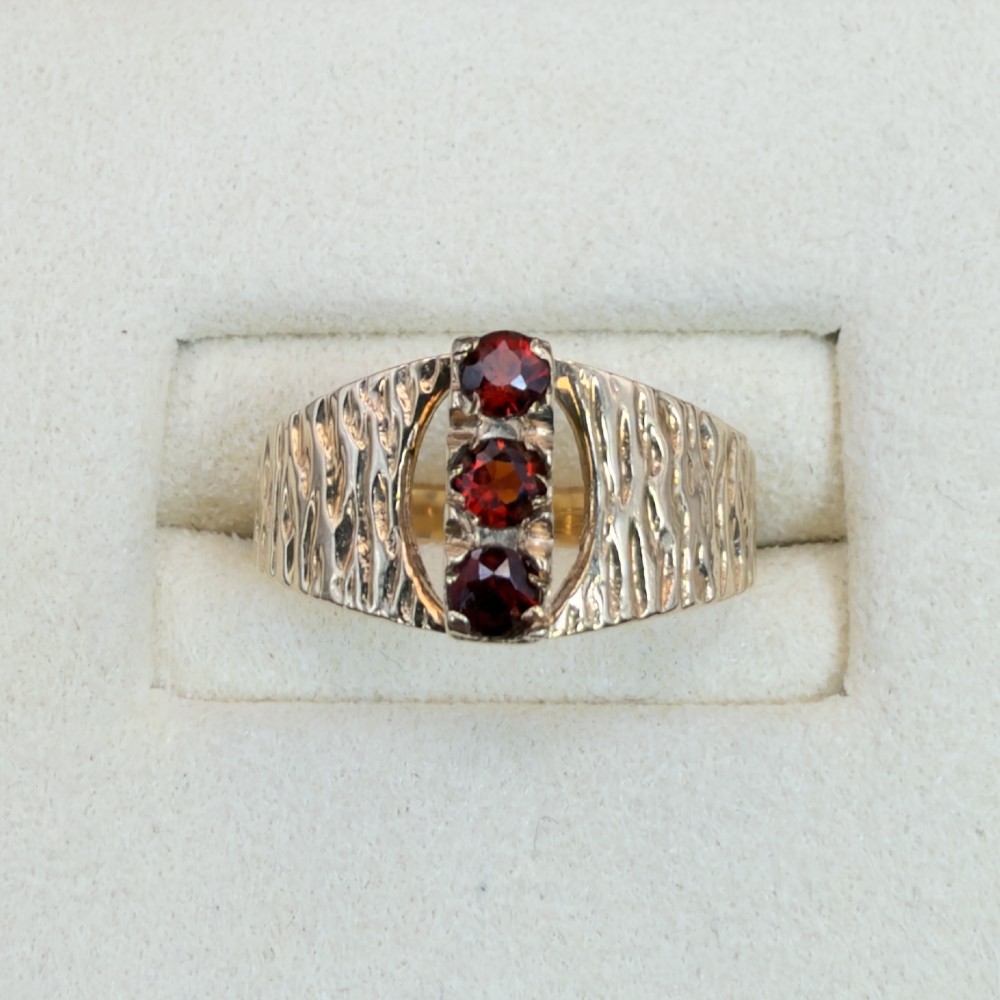 A 1960s 9ct yellow gold ring featuring a wide band with a bark effect texture and three vertically set round-cut deep red garnets. View II
