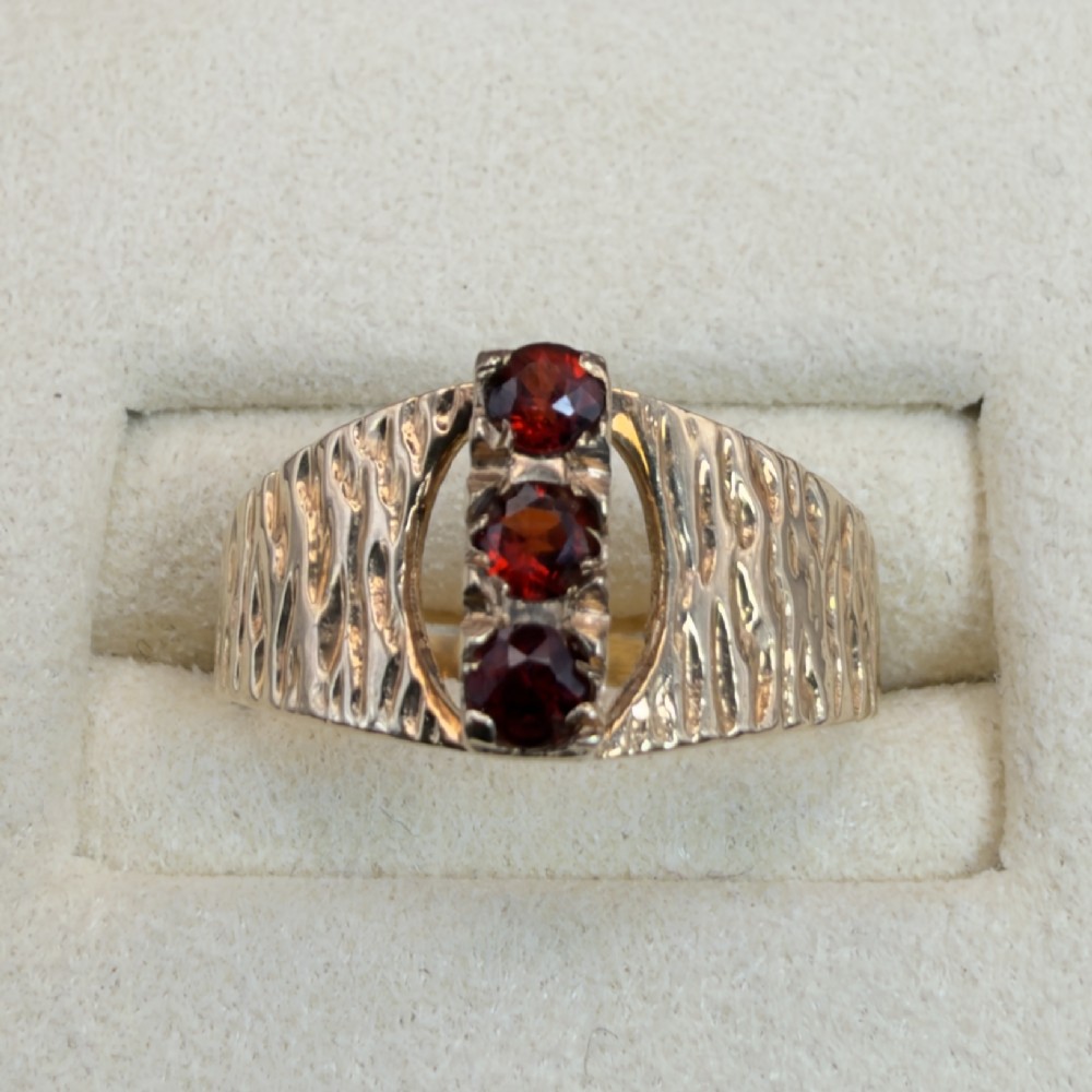 A 1960s 9ct yellow gold ring featuring a wide band with a bark effect texture and three vertically set round-cut deep red garnets. View V