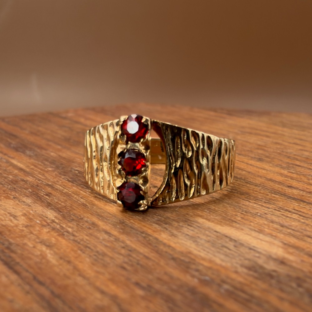 A 1960s 9ct yellow gold ring featuring a wide band with a bark effect texture and three vertically set round-cut deep red garnets. View VI