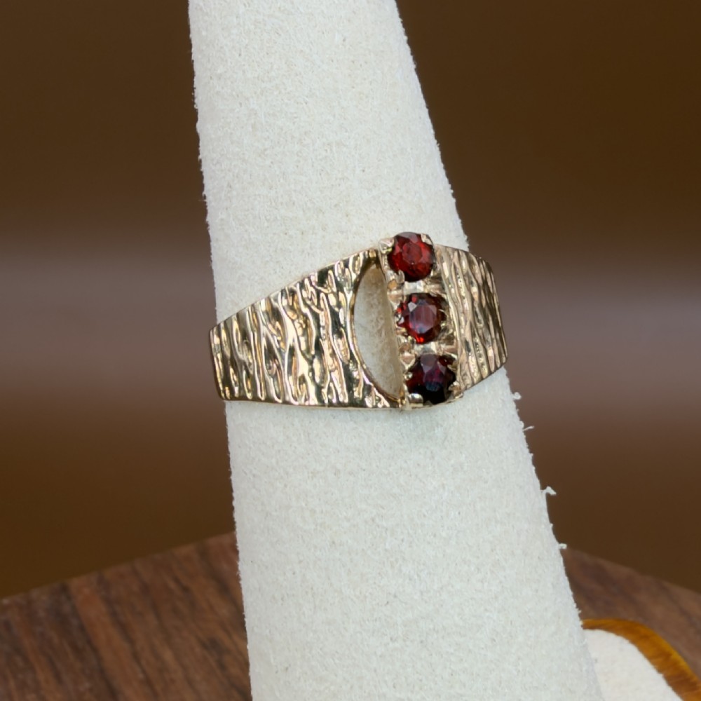 A 1960s 9ct yellow gold ring featuring a wide band with a bark effect texture and three vertically set round-cut deep red garnets. View X