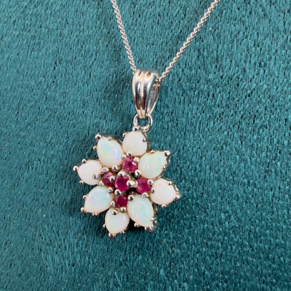 A 9ct white gold chain and cluster pendant featuring central red rubies surrounded by pear-shaped opals in a floral design. View VI