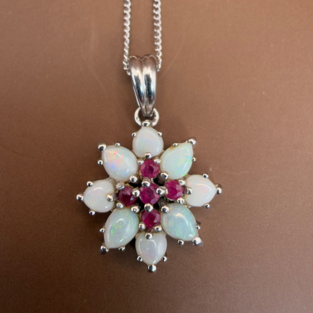 A 9ct white gold chain and cluster pendant featuring central red rubies surrounded by pear-shaped opals in a floral design. View X