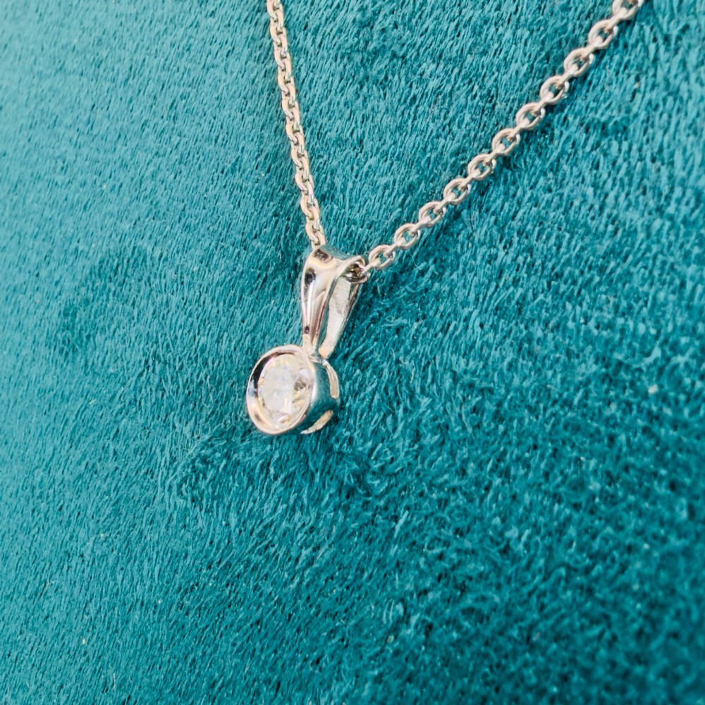 A single round brilliant-cut diamond set in a white gold bezel pendant suspended from a fine white gold chain. View IV