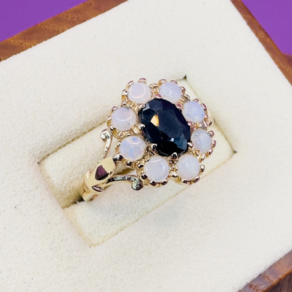 A 14ct yellow gold cluster ring featuring a central oval dark sapphire surrounded by eight white cabochon opals in claw settings. View IV