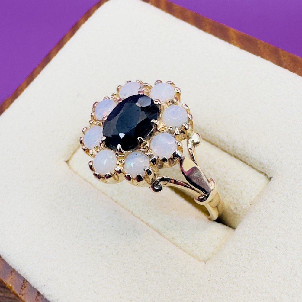 A 14ct yellow gold cluster ring featuring a central oval dark sapphire surrounded by eight white cabochon opals in claw settings. View VII