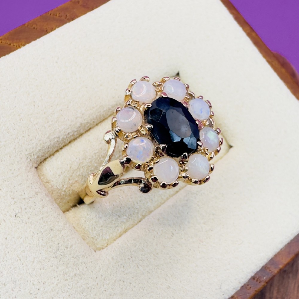 A 14ct yellow gold cluster ring featuring a central oval dark sapphire surrounded by eight white cabochon opals in claw settings. View VIII