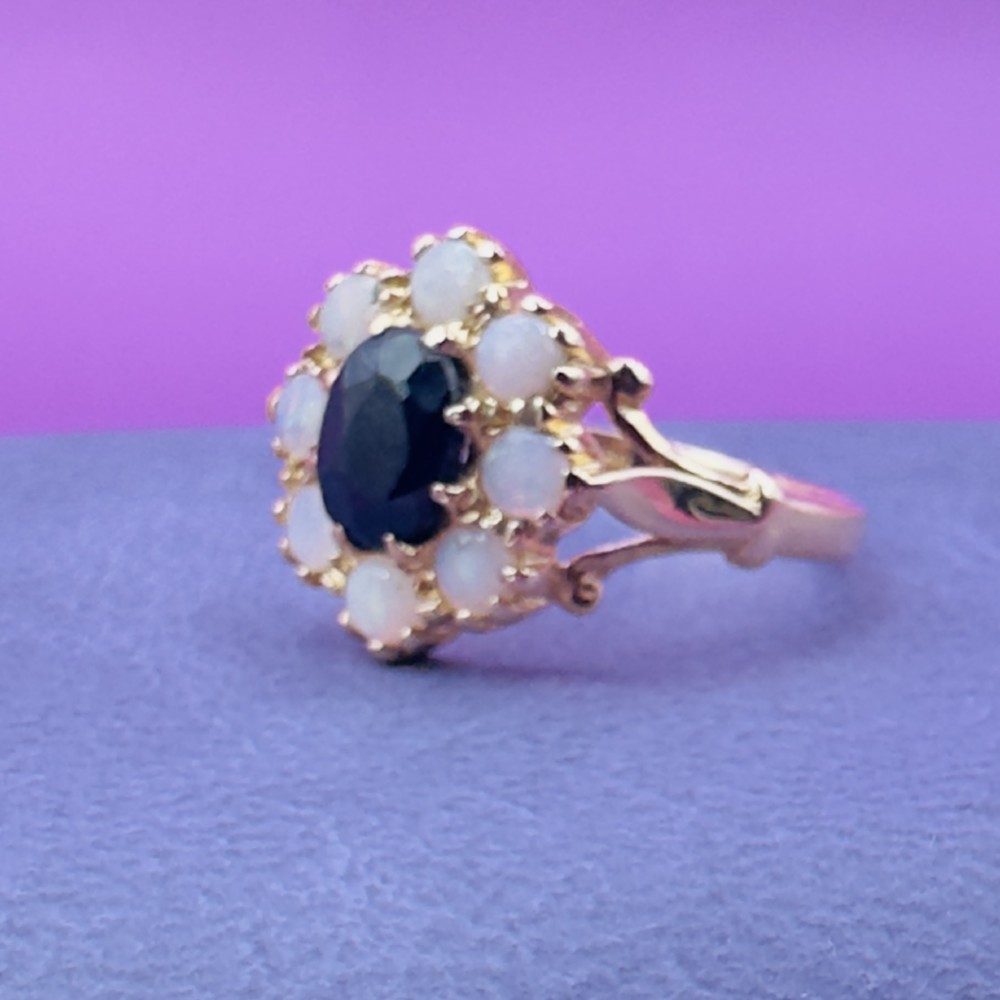 A 14ct yellow gold cluster ring featuring a central oval dark sapphire surrounded by eight white cabochon opals in claw settings. View IX