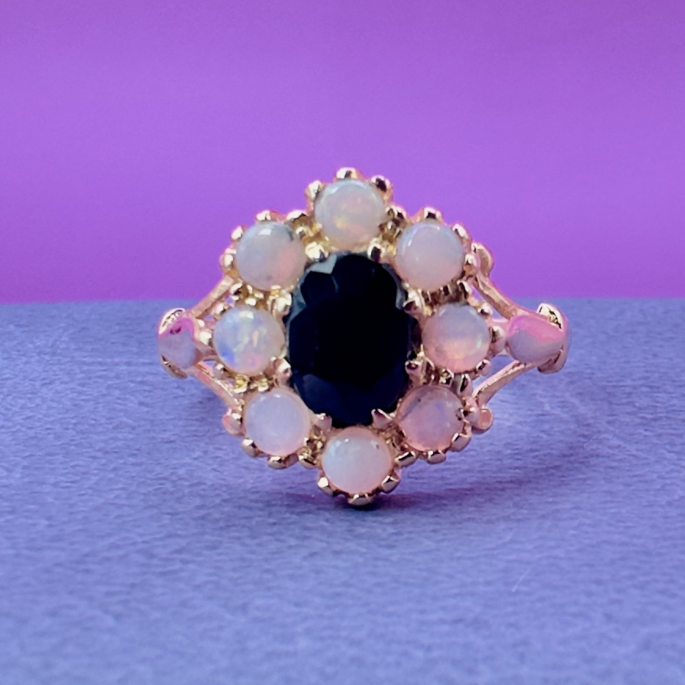 A 14ct yellow gold cluster ring featuring a central oval dark sapphire surrounded by eight white cabochon opals in claw settings. View X