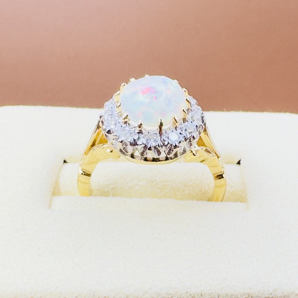 An 18ct yellow gold ring featuring a central oval cabochon opal surrounded by a halo cluster of small round brilliant cut diamonds. View V