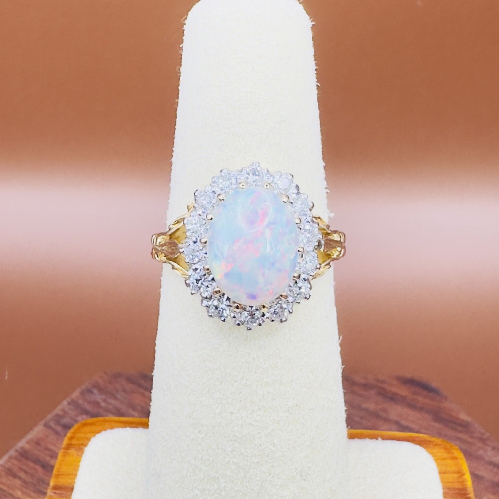 An 18ct yellow gold ring featuring a central oval cabochon opal surrounded by a halo cluster of small round brilliant cut diamonds. View VI
