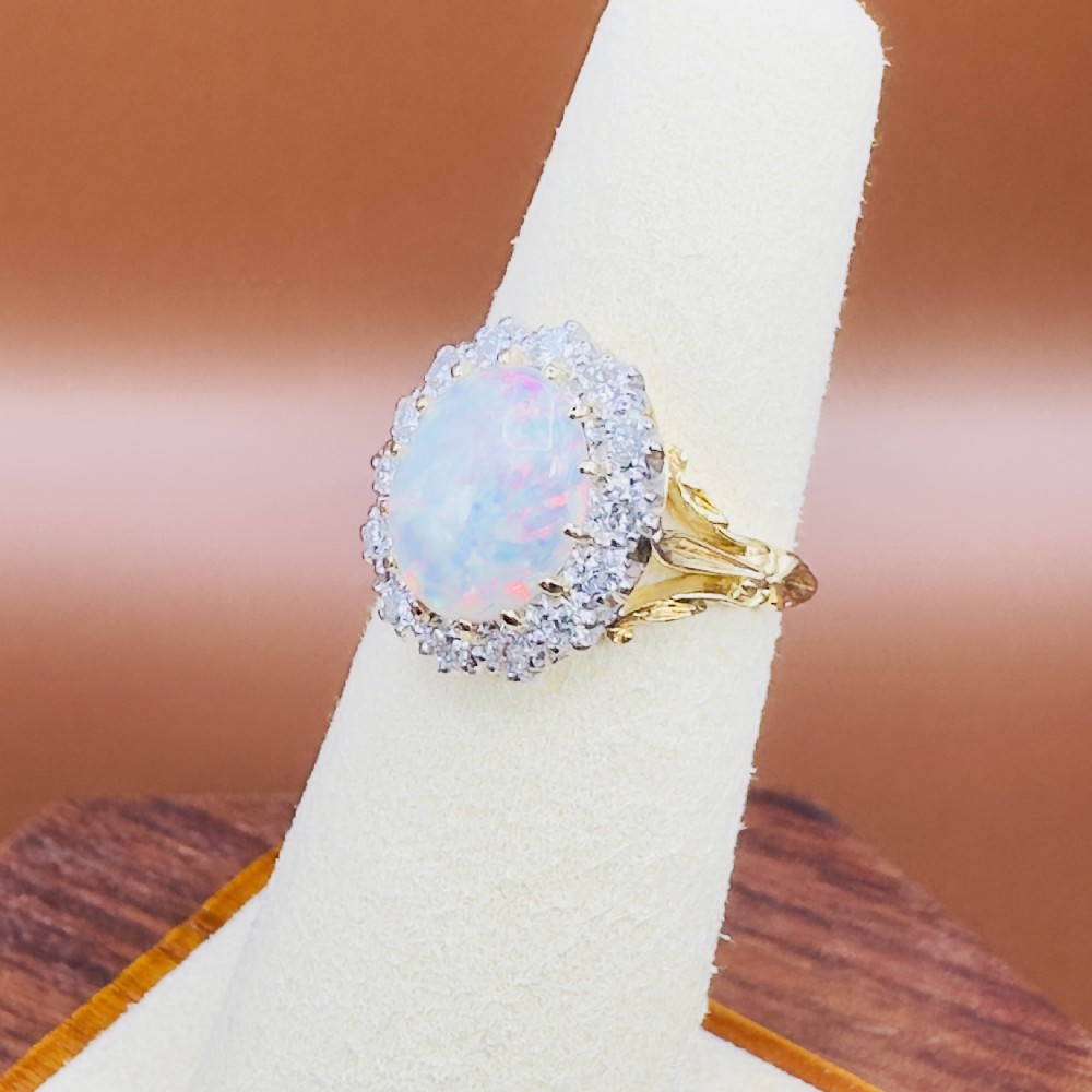 An 18ct yellow gold ring featuring a central oval cabochon opal surrounded by a halo cluster of small round brilliant cut diamonds. View VII