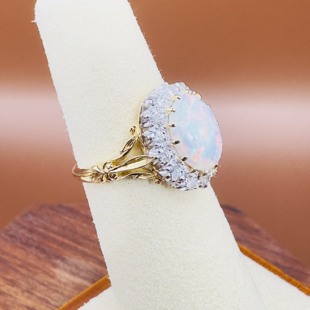 An 18ct yellow gold ring featuring a central oval cabochon opal surrounded by a halo cluster of small round brilliant cut diamonds. View VIII