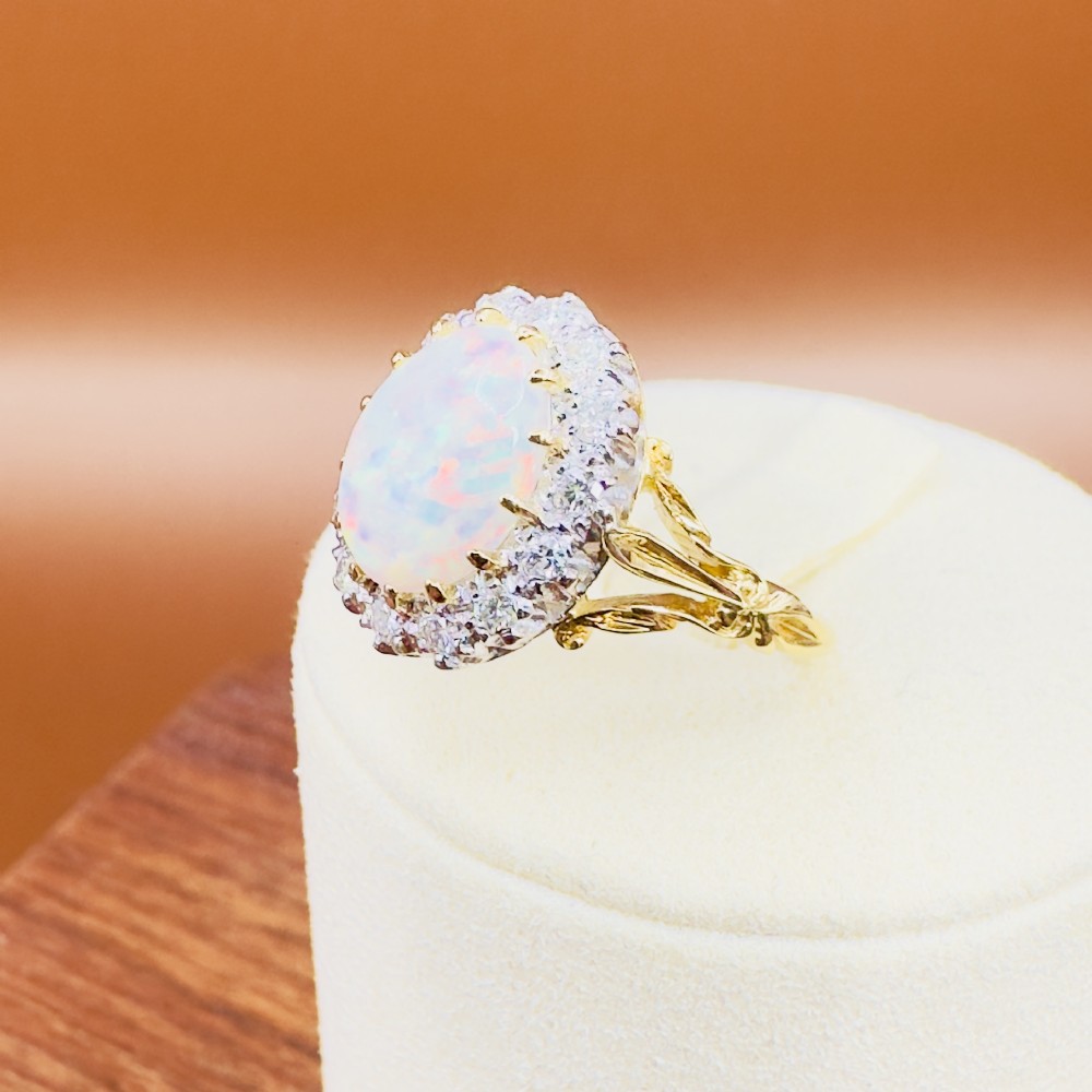 An 18ct yellow gold ring featuring a central oval cabochon opal surrounded by a halo cluster of small round brilliant cut diamonds. View IX